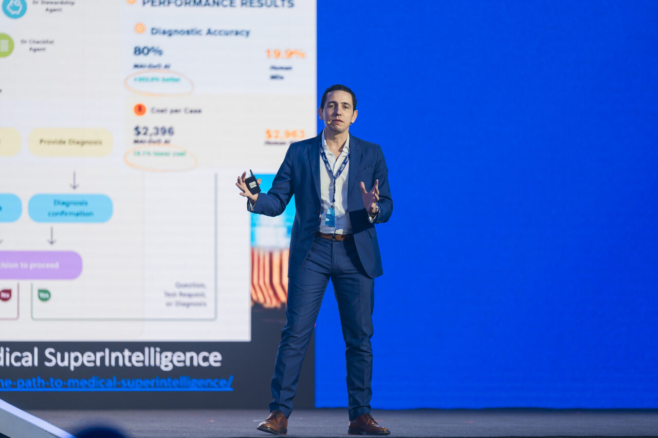 A speaker on stage at a conference, gesturing while he speaks, in front of a large screen showing performance results, with a slide comparing a diagnostic accuracy of 80% against a human baseline of 19.9% and a cost per case of $2,396 against $2,943, above the words Medical SuperIntelligence