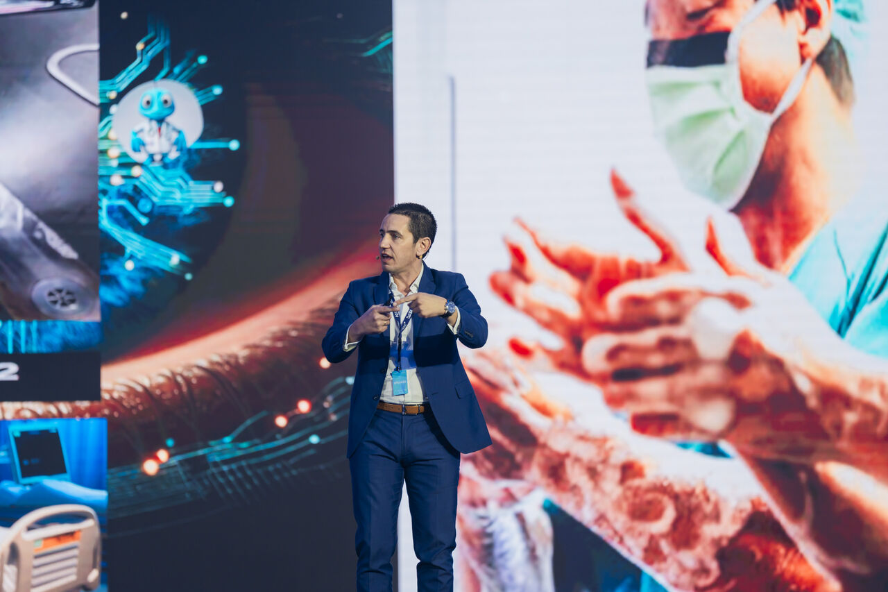 A speaker on stage at a healthcare AI keynote, gesturing as he speaks, in front of a wide screen showing an AI doctor graphic, a surgical instrument, a hospital room and a surgeon washing their hands