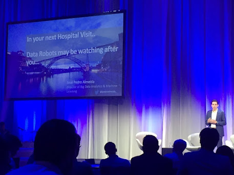 A speaker on a curtained conference stage presenting a slide that reads In your next Hospital Visit, Data Robots may be watching after you, with a seated audience in the foreground