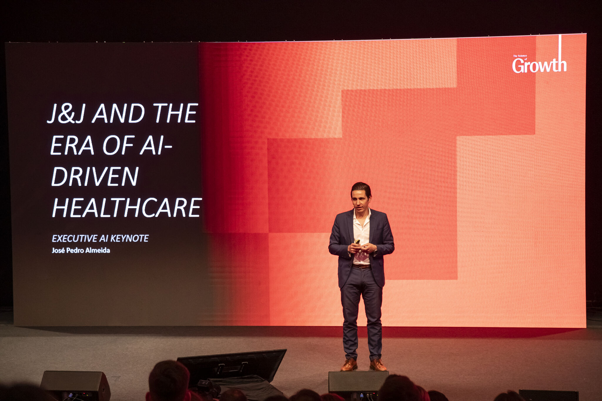 José Pedro Almeida delivering an executive AI keynote on the Johnson & Johnson stage, holding a presenter remote in front of a curved screen whose backdrop reads J&J AND THE ERA OF AI-DRIVEN HEALTHCARE, EXECUTIVE AI KEYNOTE and his name José Pedro Almeida, with stage monitors and audience heads in the foreground