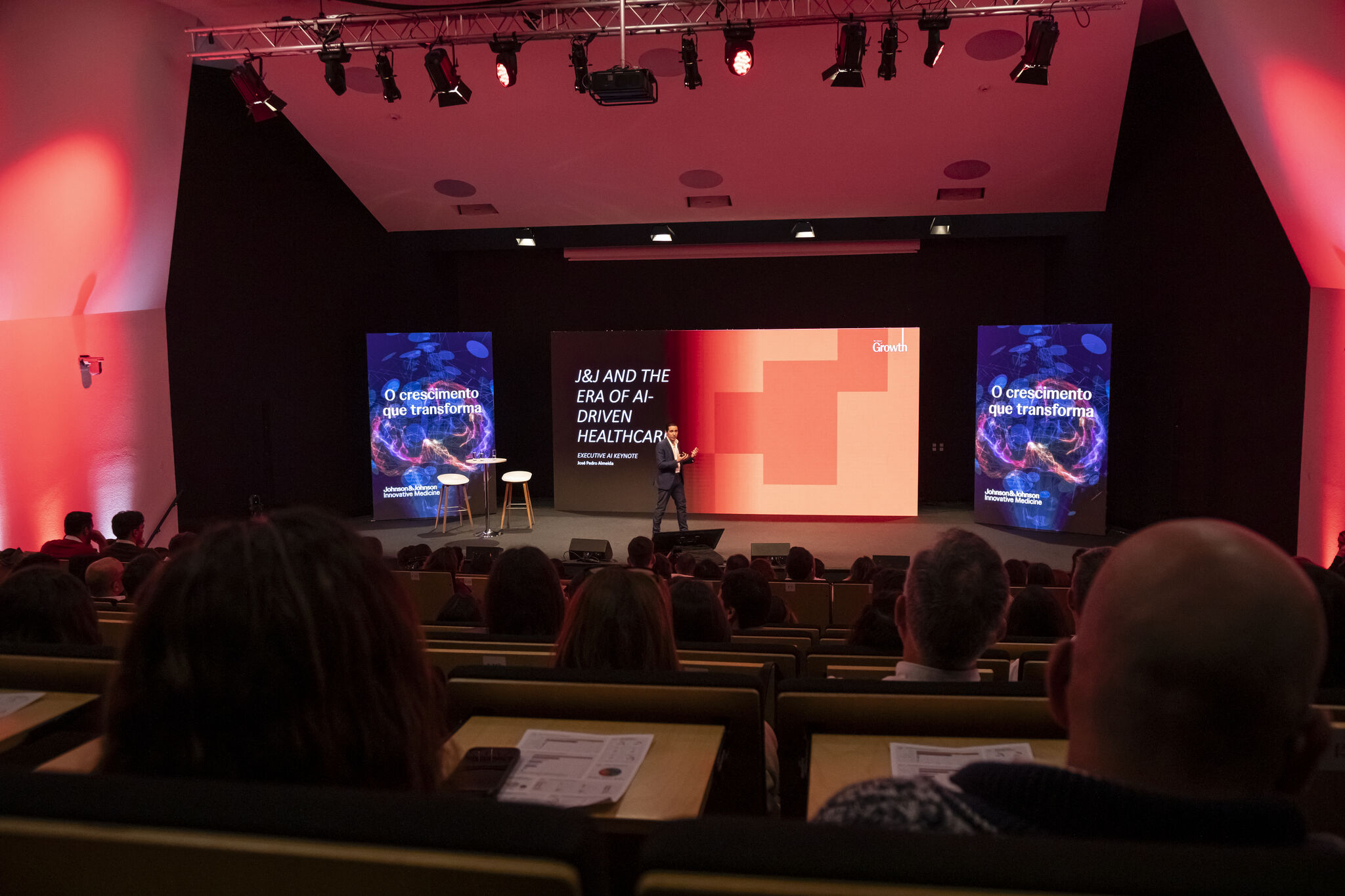 A speaker on stage in an auditorium in front of a screen reading J&J AND THE ERA OF AI-DRIVEN HEALTHCARE and EXECUTIVE AI KEYNOTE, flanked by banners reading O crescimento que transforma and the Johnson & Johnson Innovative Medicine logo, with the audience seated in the foreground