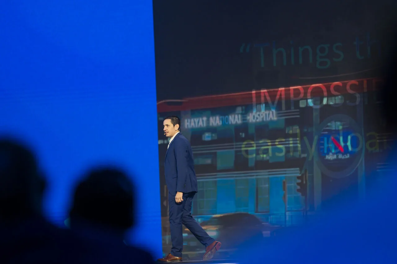 José Pedro Almeida on stage at a healthcare AI event in front of a screen showing Hayat National Hospital with the word Impossible, audience silhouettes in the foreground