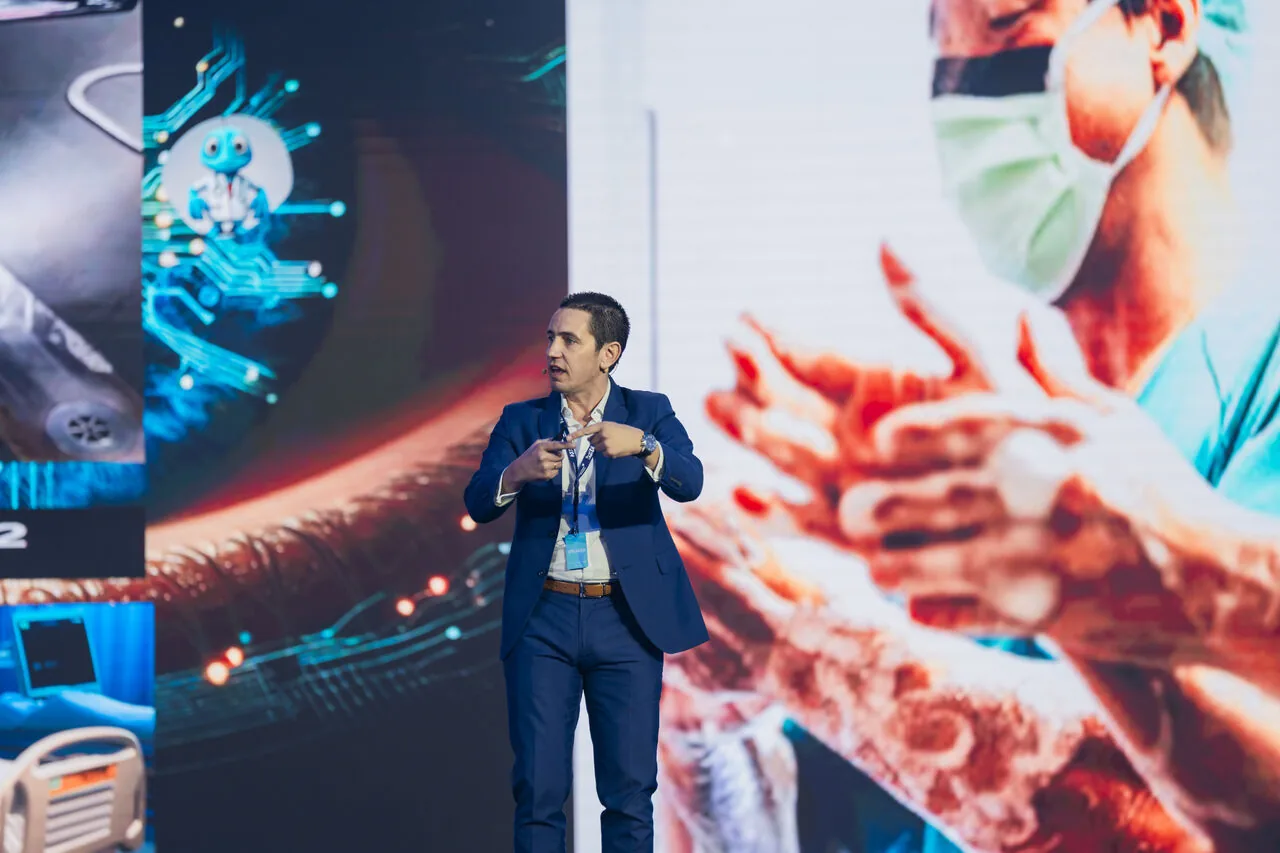 José Pedro Almeida presenting on stage at a healthcare AI event, with screens showing a robot, medical equipment and a gloved clinician